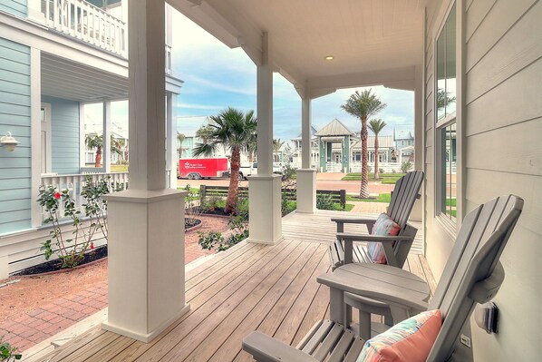 A covered porch with wooden flooring, a seating area with chairs and cushions, and a view of the street with palm trees and a red delivery truck. - Sunset Regatta 230 GR (Port Aransas)