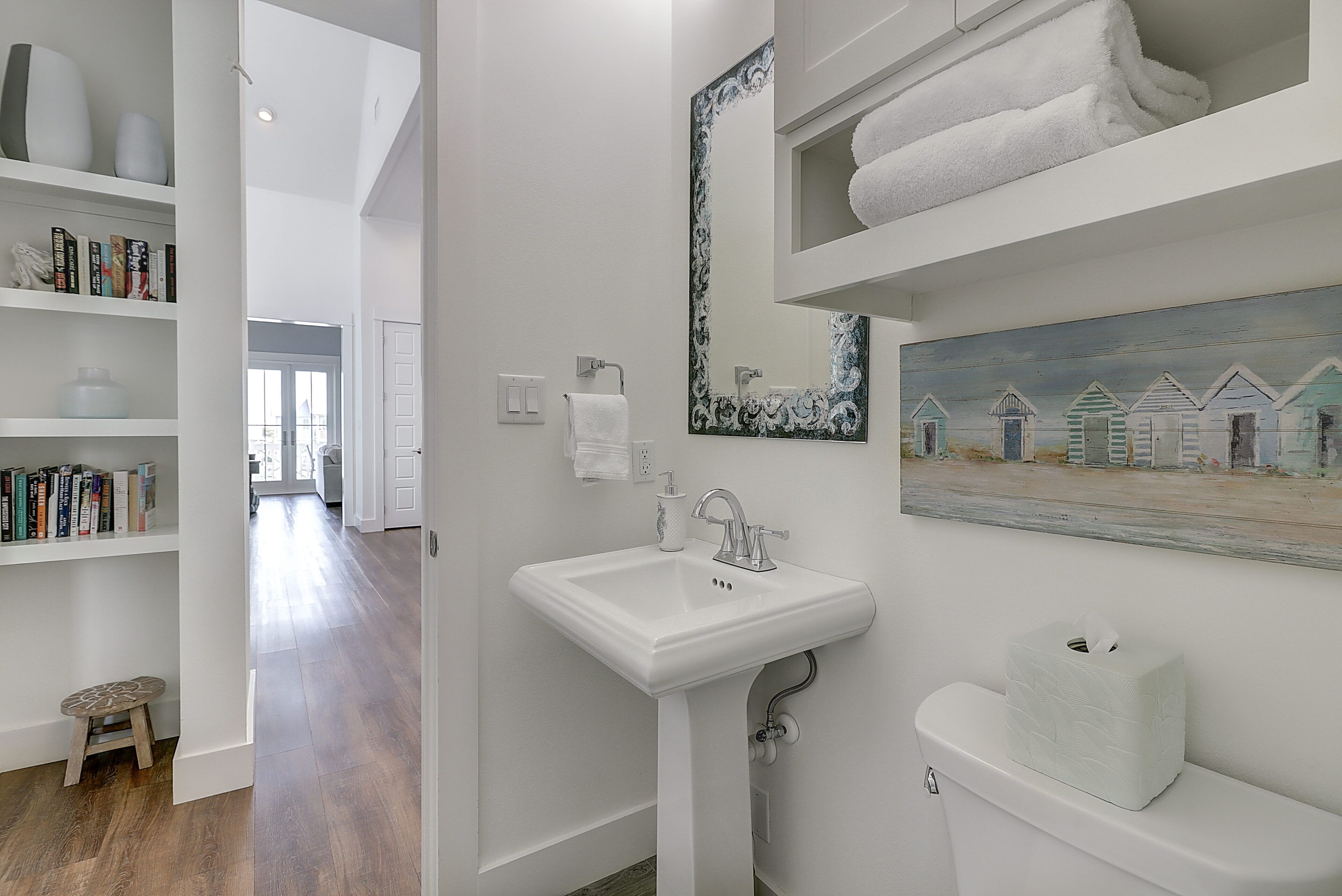 A bathroom with a white sink, a mirror, and a painting of beach huts. There are shelves with books and a toilet paper holder.