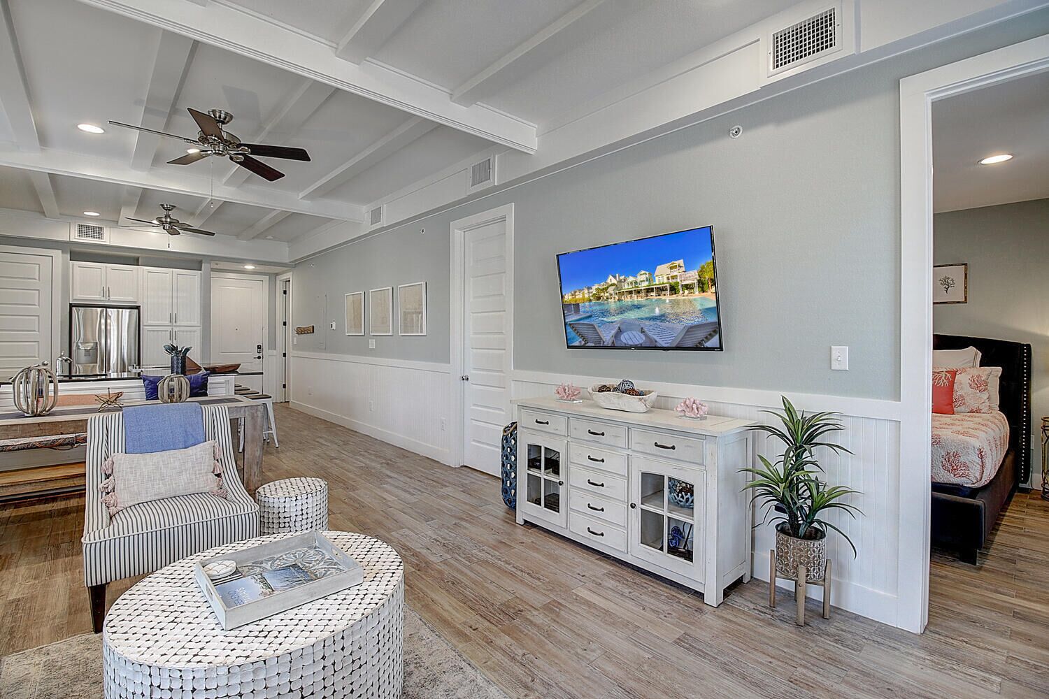 A spacious living area with a large flat-screen TV mounted on the wall, a dining area with white cabinetry, and a cozy seating area with a round wicker table and striped chair.