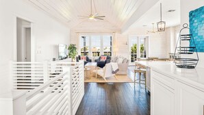 A spacious living area with a high ceiling, a ceiling fan, and a large window providing natural light. There is a seating area with a sofa and chairs, and a kitchen area with a white countertop and a hanging light fixture. - The Hideaway 249 HI (Port Aransas)