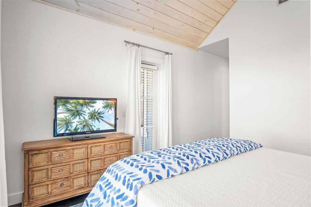 Un dormitorio con cama, cómoda de madera, televisor y ventana con cortinas blancas.