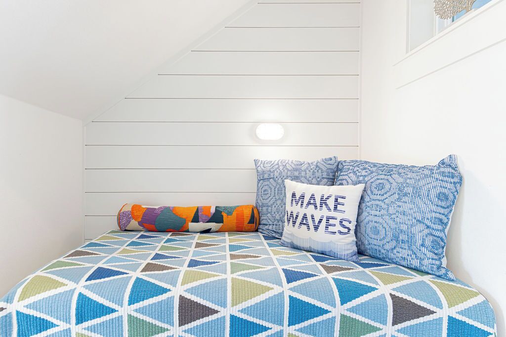 A bedroom with a geometric-patterned bedspread, a pillow with "MAKE WAVES" text, and a colorful rolled blanket.