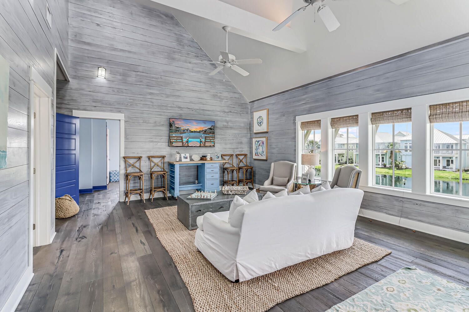 A spacious living room with a high ceiling, wooden walls, and large windows offering a view of a house and a lake.