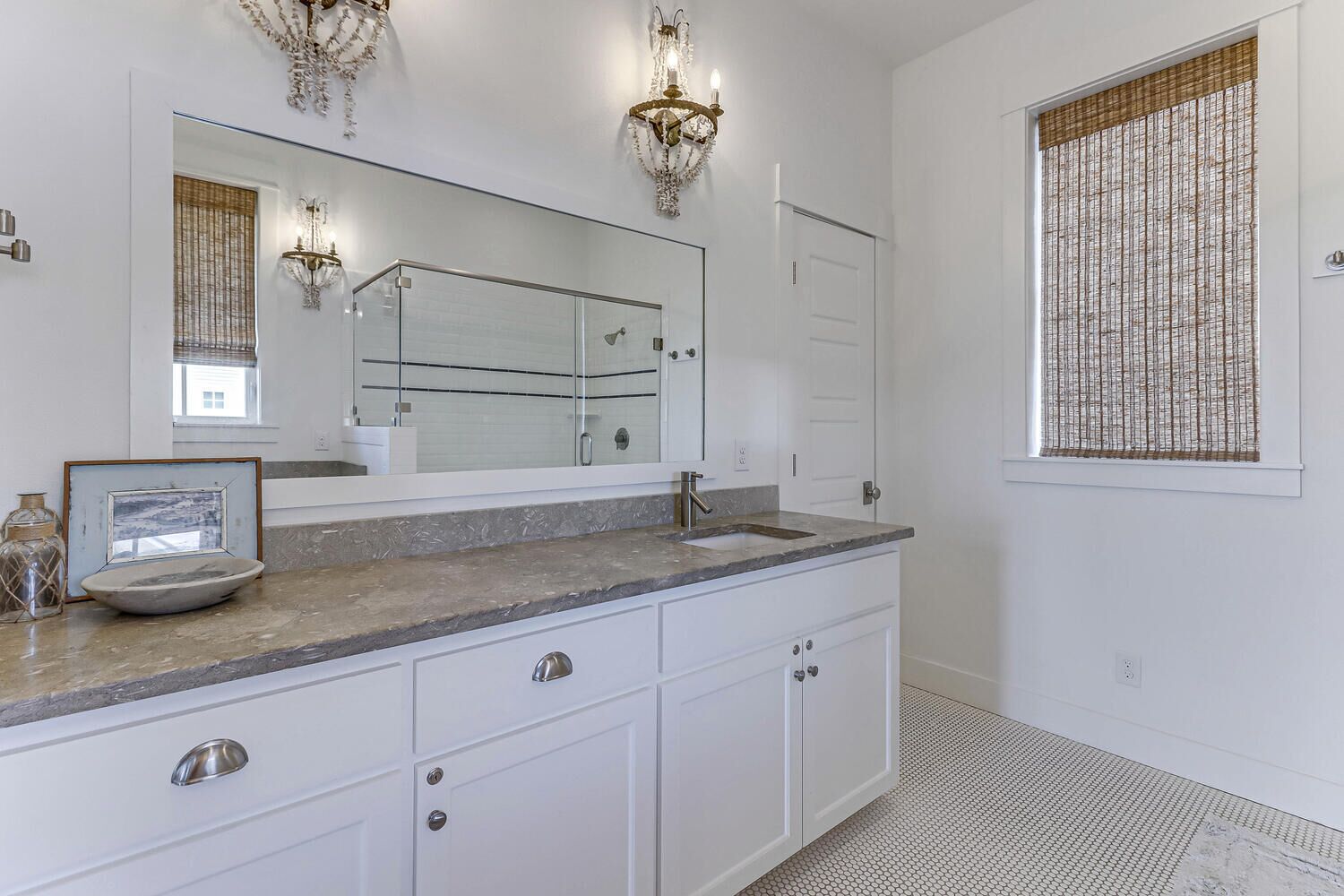 A bathroom with a large mirror, two wall sconces, a sink, and a window with blinds.