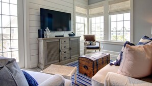A living room with a television, a wooden cabinet, a cushioned chair, a wooden chest, and a striped rug. - So Long TC 10305 (Port Aransas)