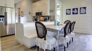 A modern kitchen with white cabinetry, a stainless steel refrigerator, a microwave, and a dining area with a white table and four chairs. - So Long TC 10305 (Port Aransas)