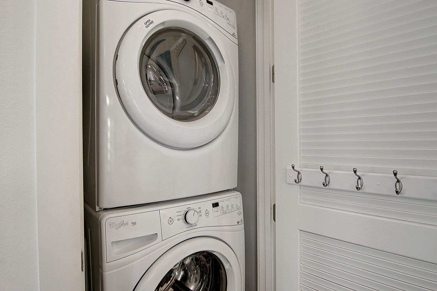 A stacked washer and dryer unit in a laundry room with a white door and a metal hook system on the wall.