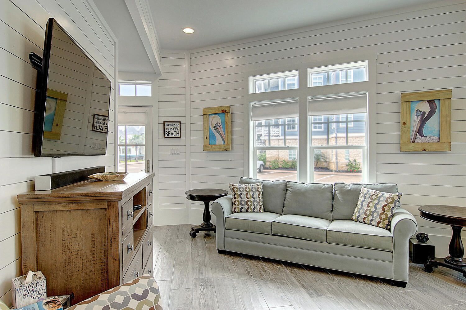 A room with a wooden cabinet, a gray sofa with patterned cushions, a round table, and a large window with multiple panes.