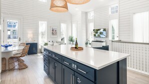 A spacious kitchen with a large island, white wooden paneling, and wicker chairs.
