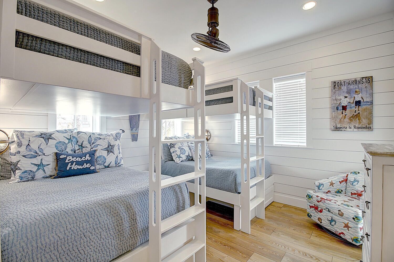Une chambre avec des lits superposés, un lit agrémenté d’un coussin sur le thème de la plage, un mur orné d’un tableau et un ventilateur de plafond.