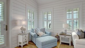 Une pièce avec un canapé bleu clair, un fauteuil blanc et une petite table avec une lampe. On y trouve des fenêtres avec des volets, un plancher en bois et une porte blanche.