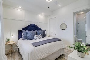 A bedroom with a large bed, a bedside table with a lamp, a mirror on the wall, and a bathroom with a shower and sink.