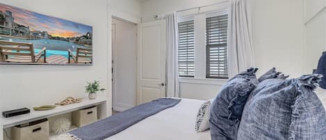 Une chambre à coucher avec un lit, un tableau représentant un paysage côtier, un bureau avec une plante et une fenêtre avec des stores.