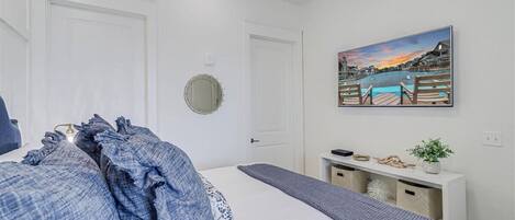 A bedroom with a bed, a bedside table, a mirror, and a painting of a waterfront scene.
