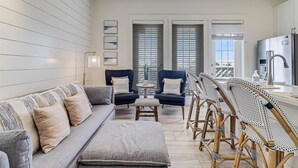 Ein modernes Wohnzimmer mit einem grauen Sofa, zwei blauen Sesseln, einem kleinen runden Tisch und einer Küchenzeile mit Kühlschrank und Spüle.