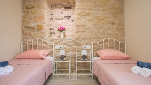 Une chambre avec deux lits, chacun recouvert d’une couvre-lit rose et agrémenté d’oreillers assortis, un mur en pierre et une petite étagère sur laquelle est posé un vase de fleurs.