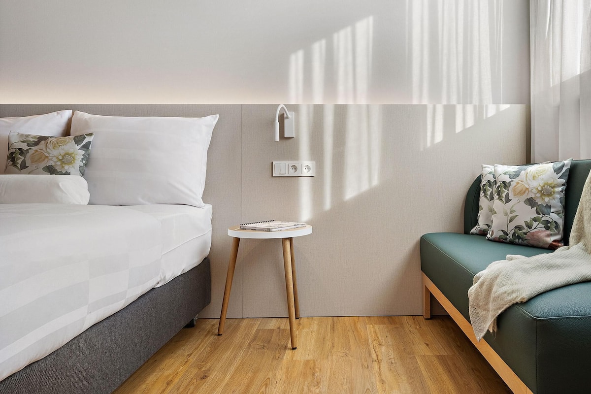 Quarto de hotel moderno com uma cama, uma mesinha de apoio, um sofá e um aplique na parede.