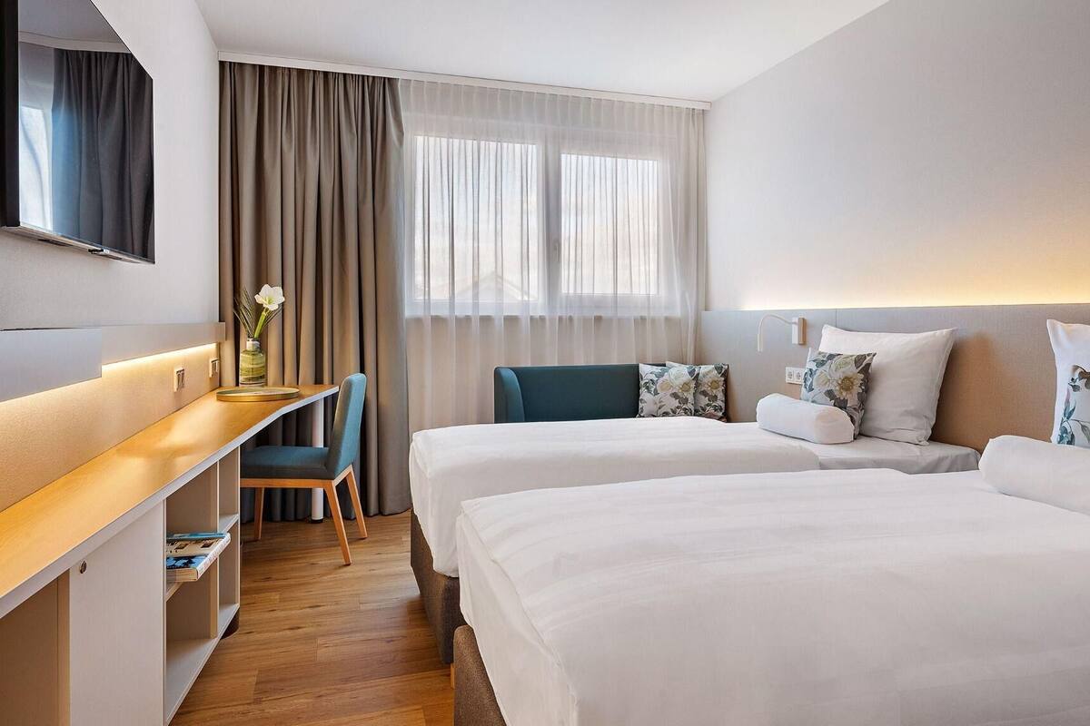 A hotel room with two beds, a desk, a chair, and a large window with curtains.