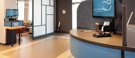 A modern reception desk with a digital screen displaying text, a coffee machine, and seating area with chairs and tables.