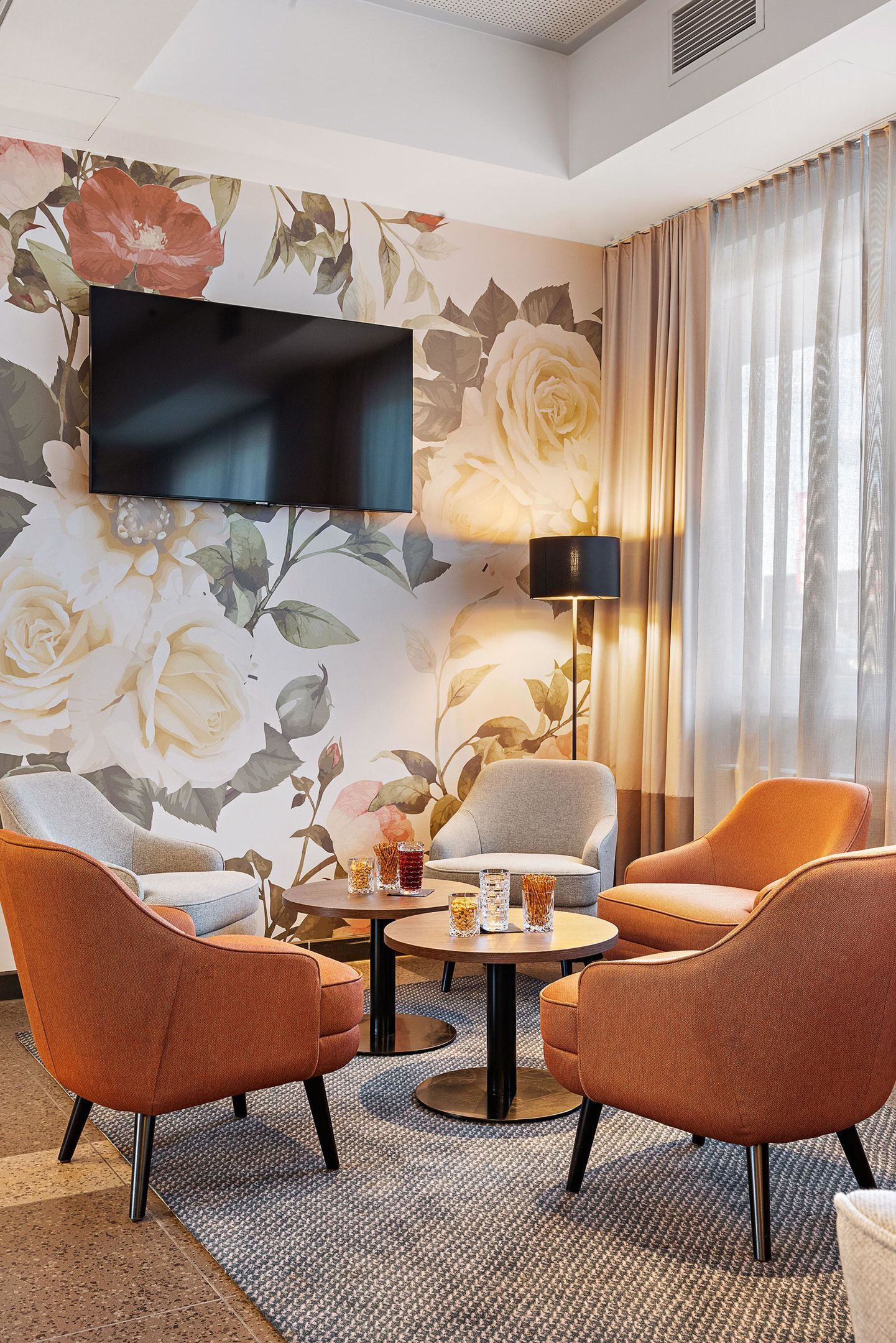 Une chambre d’hôtel dotée d’un téléviseur à écran plat, d’un papier peint à motifs floraux, de deux fauteuils orange, d’une table ronde avec des boissons et d’un lampadaire.