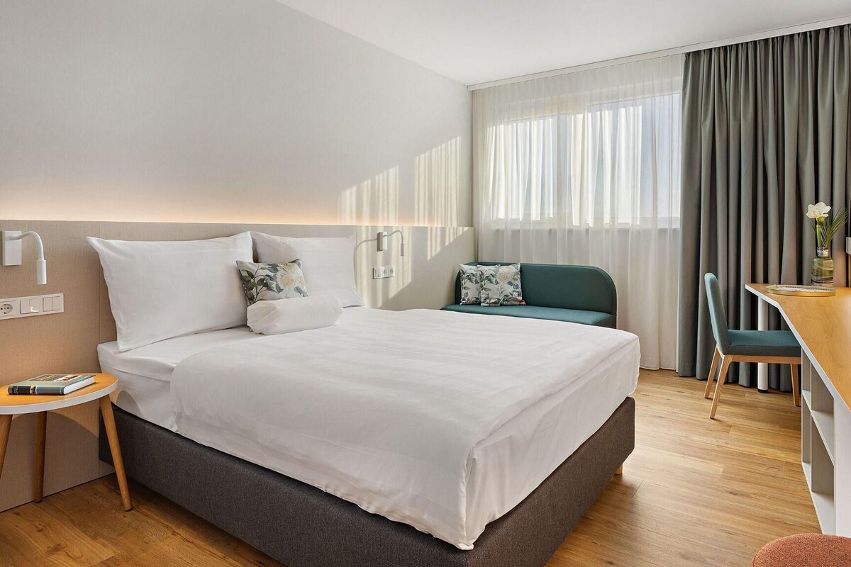 A hotel room with a bed, bedside table, chair, desk, and curtains.