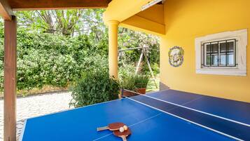 A blue ping pong table with paddles and balls, a yellow wall with a window, and a wooden pergola structure.