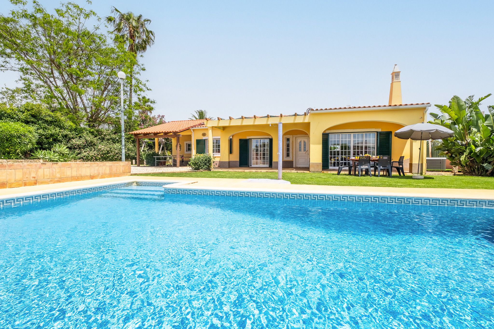 Una propiedad residencial con piscina, un patio con mobiliario exterior y una casa amarilla con techo de tejas.