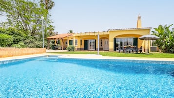 Una propiedad residencial con piscina, un patio con mobiliario exterior y una casa amarilla con techo de tejas.