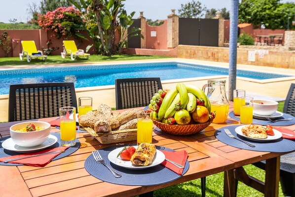 A dining table set with breakfast items, a fruit bowl, and a view of a swimming pool.