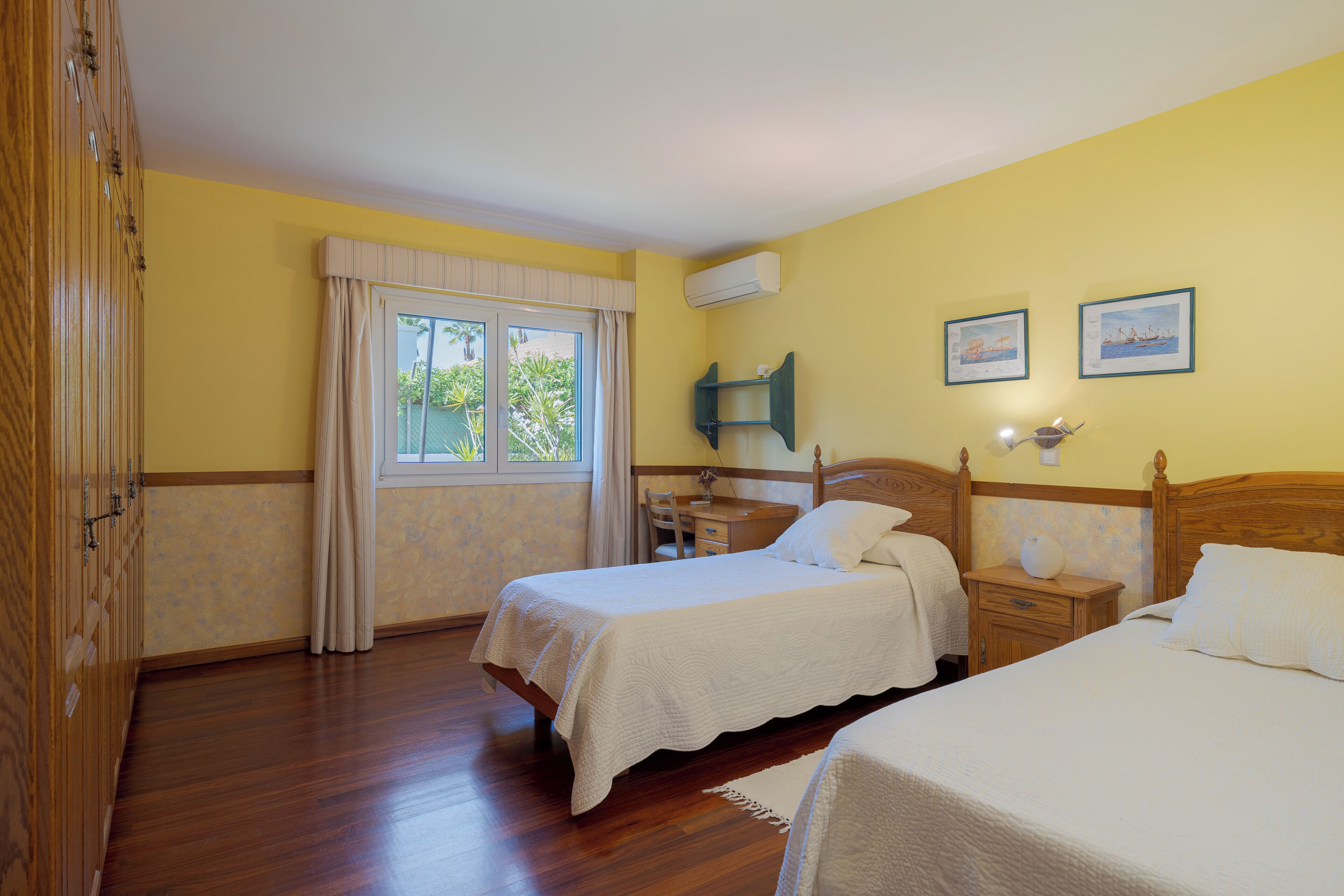 Une chambre avec deux lits simples, une armoire en bois, une fenêtre avec des rideaux, une table de chevet avec une lampe et deux tableaux encadrés accrochés au mur.