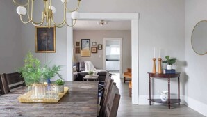 A dining area with a wooden table, chairs, and a chandelier. There is a side table with candles and a plant. The room has a neutral color scheme with white walls and a gray ceiling.