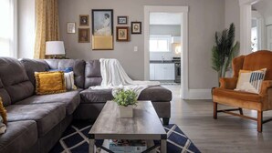 A living room with a sectional sofa, a coffee table, and a chair. There is a kitchen visible through an open doorway.