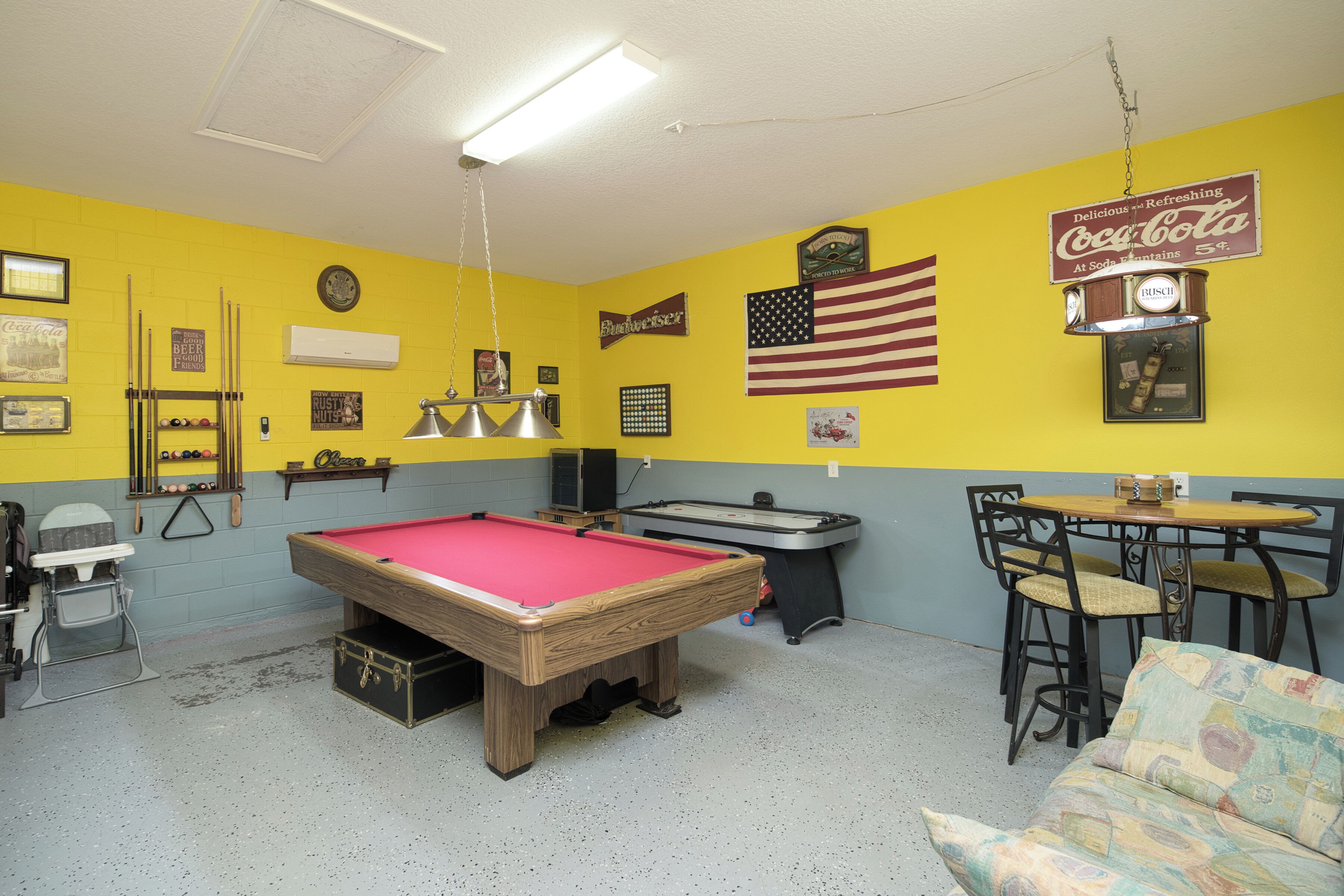 Une table de billard dans une pièce dont les murs sont peints en jaune et en bleu, avec un drapeau américain et une enseigne Coca-Cola.