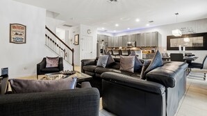 A modern living room with a black leather sofa, a glass coffee table, and a dining area with a kitchen in the background. - Blissful Retreat 6 Bedrooms (Davenport)