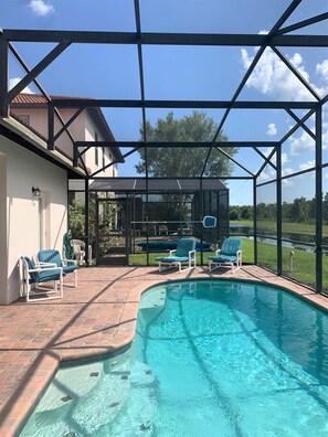 Une piscine extérieure couverte, dotée de chaises longues, sous un ciel bleu azur, avec une maison visible au loin.