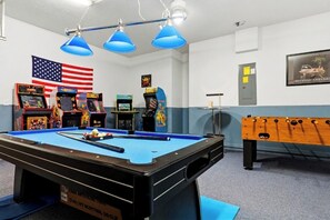 A pool table with a blue felt surface, a foosball table, and arcade games in a room with white walls and a blue accent wall.