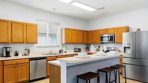 A modern kitchen with wooden cabinets, stainless steel appliances, a central island with stools, and a window with blinds.
