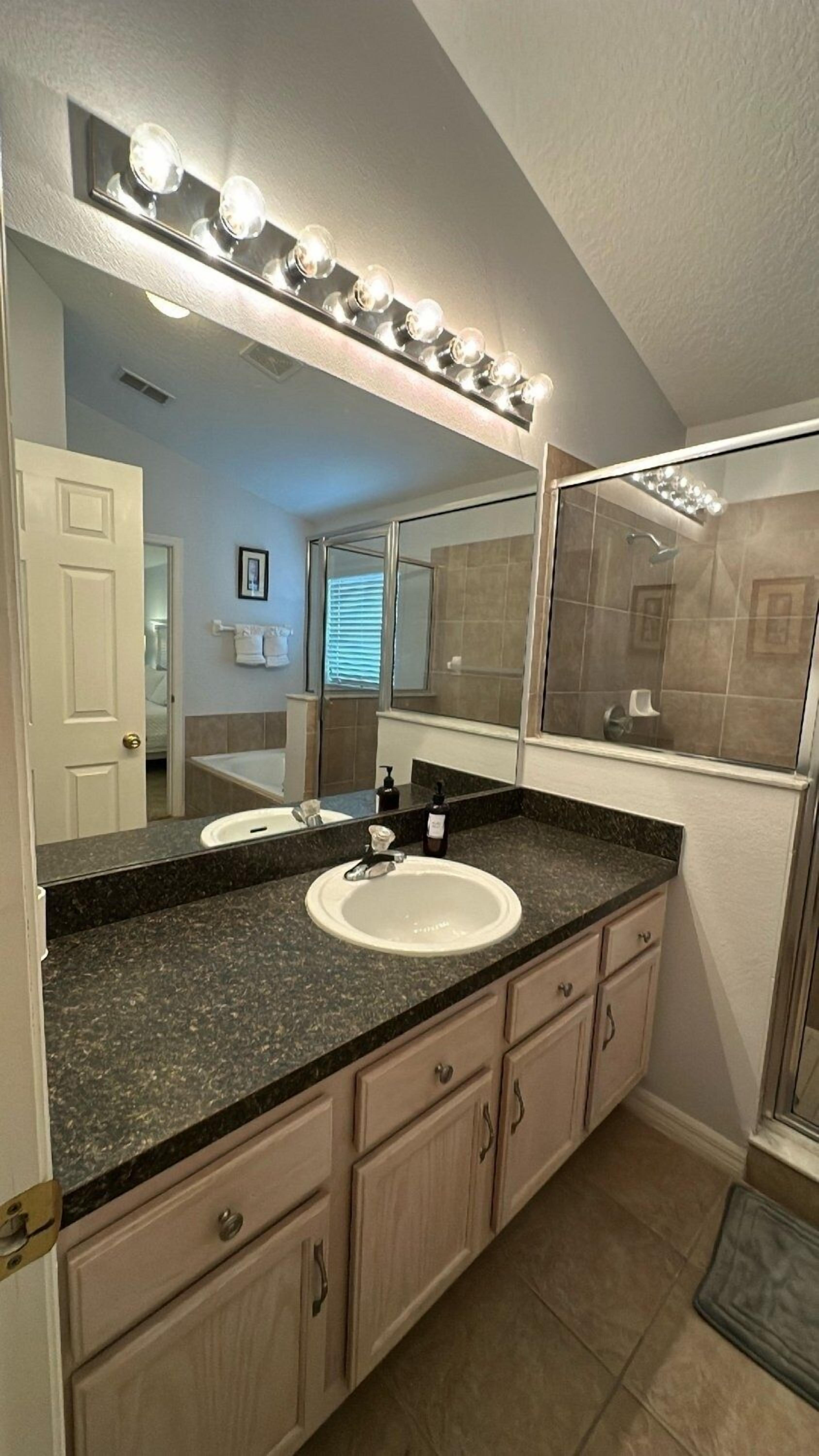 A bathroom with a double sink vanity, a large mirror with lights, a shower, and a door leading to another room.
