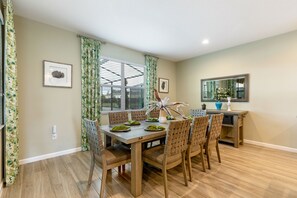 Un coin repas avec une table et des chaises en bois, une fenêtre avec des stores et une petite table d’appoint sur laquelle est posé un vase.