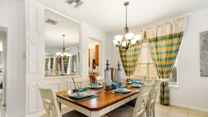 Comedor con mesa de madera dispuesta para cuatro personas, sillas blancas, una araña y cortinas a rayas.