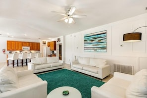 A modern living room with white sofas, a green rug, and a kitchen in the background.