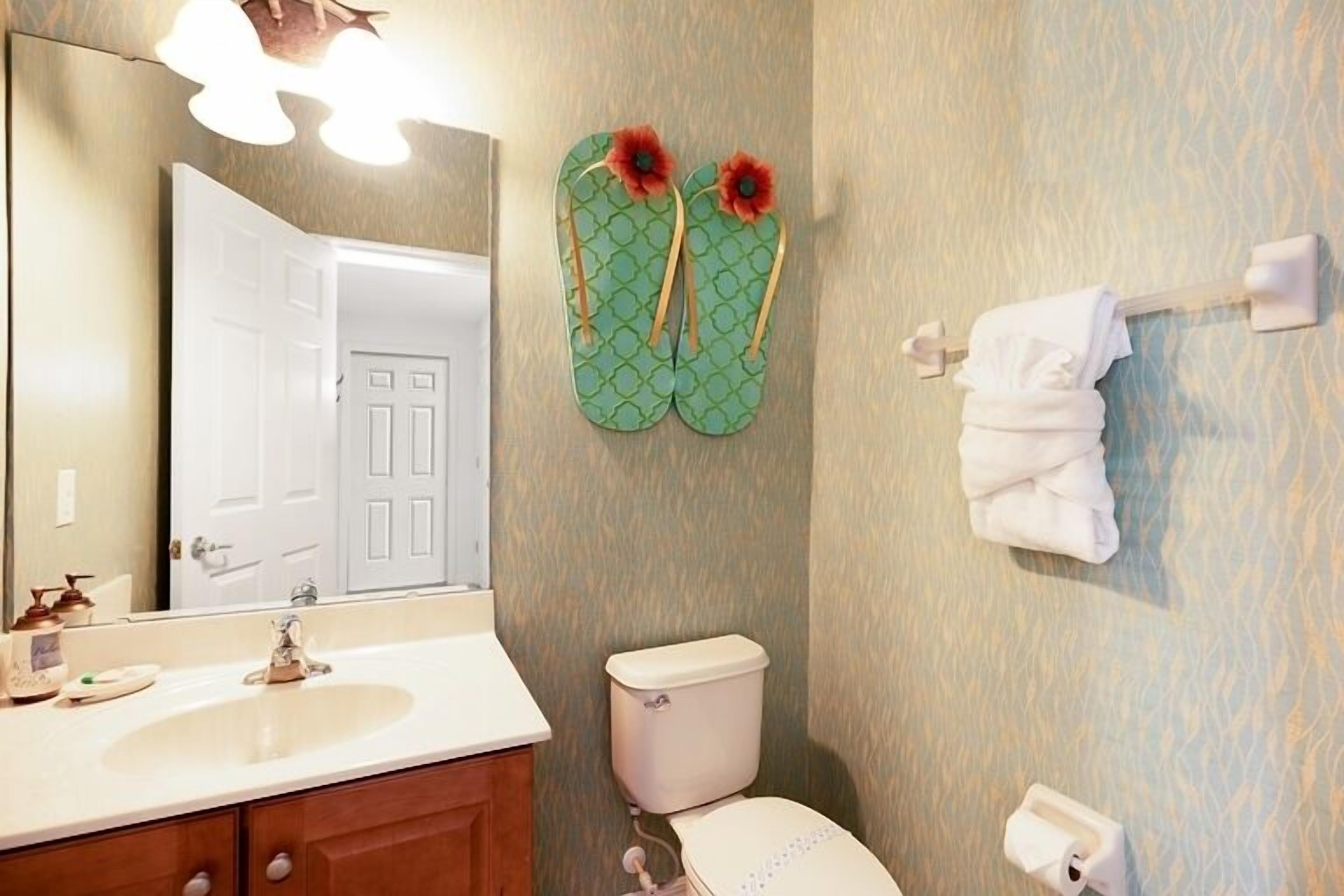 Ein Badezimmer mit Waschbecken, Toilette und Spiegel. Grüne Flip-Flops hängen an der Wand, und ein Handtuchhalter mit einem gefalteten Handtuch ist vorhanden.