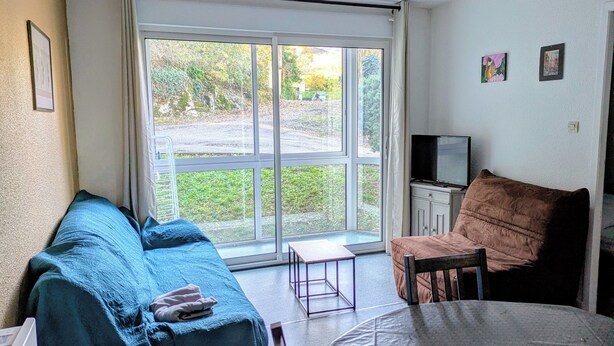 Una habitación con un ventanal grande, un sofá, una silla, una mesita y un televisor.
