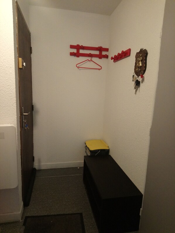 A small, dimly lit room with a door, a coat rack with hangers, a small bench, and a wall-mounted coat hook.