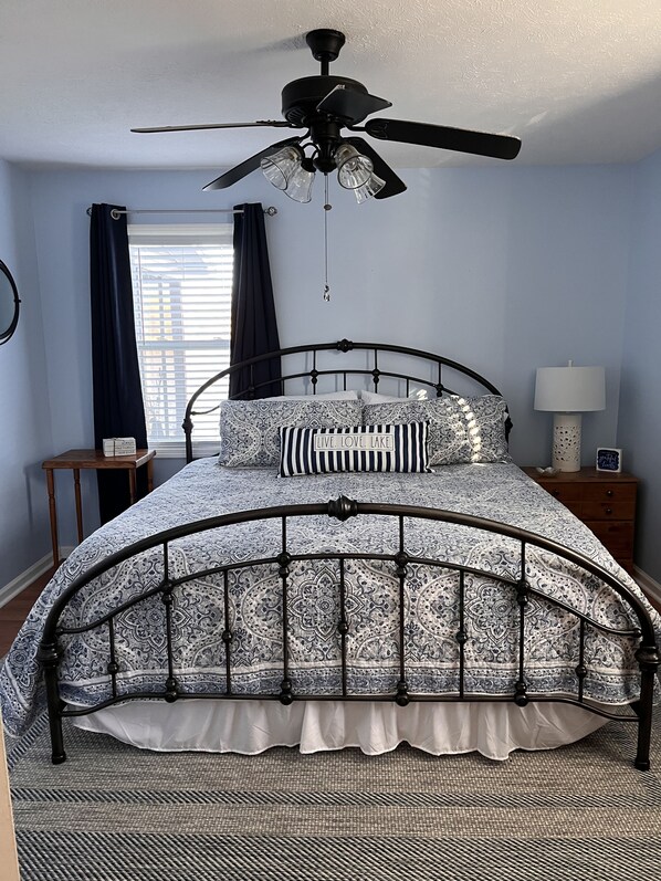 Un dormitorio con cama, ventilador de techo, ventana con cortinas, mesita de noche y velador.