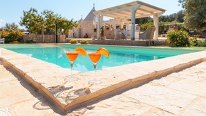 A pool area with two cocktails on a stone ledge, a covered seating area, and a stone building in the background.