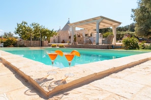 A pool area with two cocktails on a stone ledge, a covered seating area, and a stone building in the background.