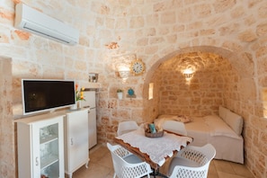 A stone-walled room with a television on a cabinet, a dining table with chairs, and a cozy seating area with cushions.