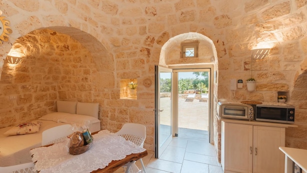 A stone-walled room with a bed, a table with a lace tablecloth, a basket of bread, and a kitchen area with a microwave.