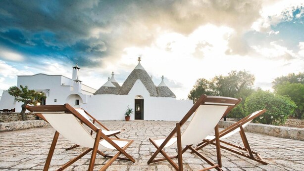 A white building with a unique architectural style, featuring multiple conical roofs and a central door. In front of the building, there are two wooden lounge chairs positioned on a cobblestone patio. - Trullo Paleo (Ceglie Messapica)